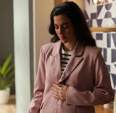 Mulher em um blazer rosa segurando a barriga, em pé em um ambiente fechado.